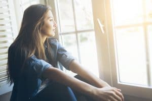 Smiling young woman sitting and looking through the window. With long hair, wears denim shirt. Hands on legs.